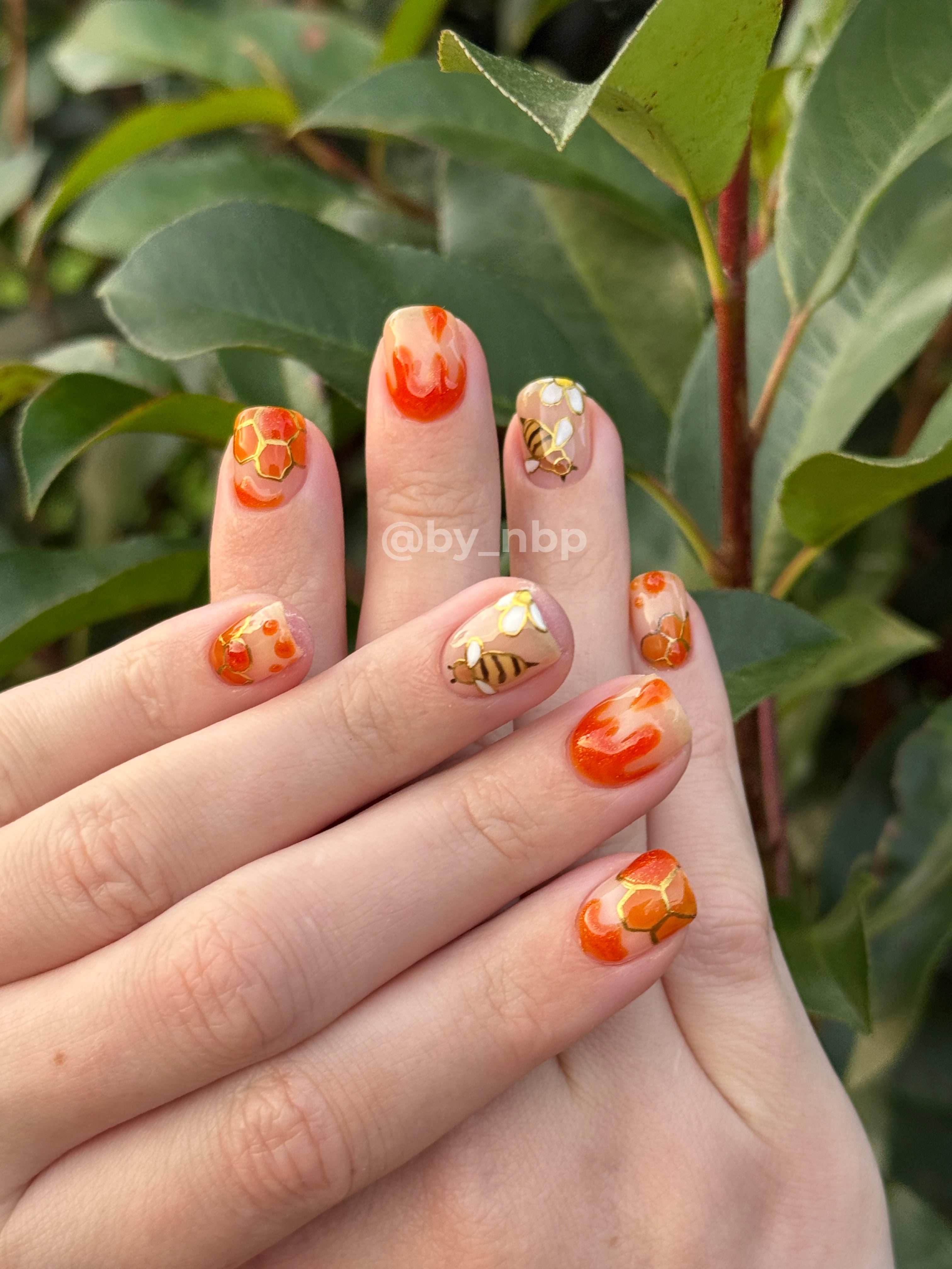 Client hand with short squoval shaped builder gel (BIAB) nails with glossy orange & gold bee/honey freestyle nail art.